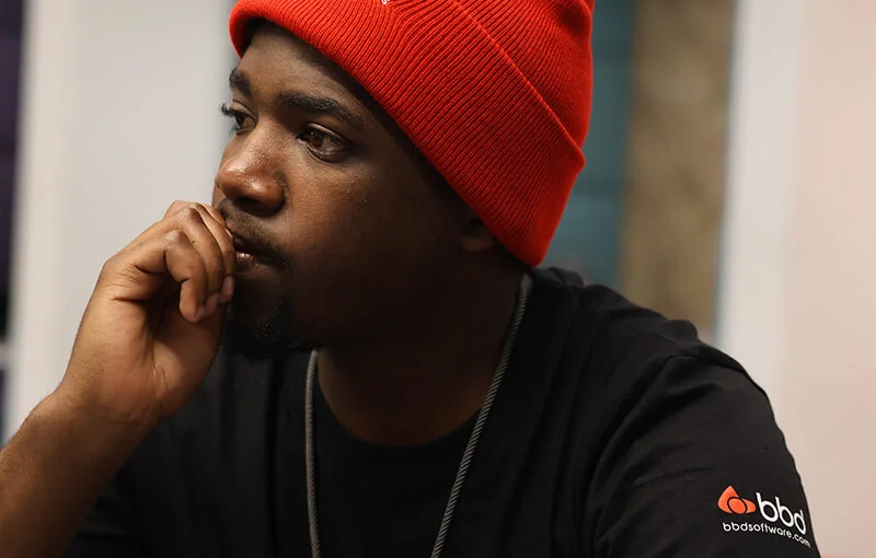 A man wearing a red beanie and black t-shirt with a logo sits pensively, resting his chin on his hand, conveying a thoughtful and focused mood.