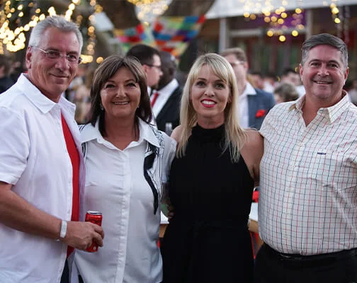 A group of four people smiling at an outdoor event with festive lights and a colorful banner in the background. They appear relaxed and joyful.