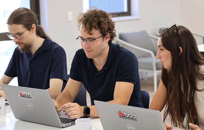 Three people sit at a table working on laptops, engaged in focused discussion in a bright office setting. They exude concentration and teamwork.