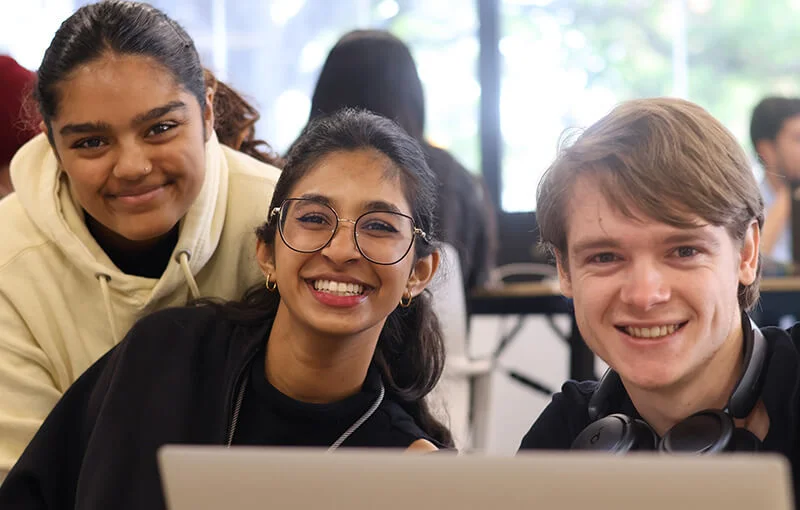 Three young people smile warmly at the camera in a classroom setting. A laptop is visible in the foreground, suggesting a collaborative study session.