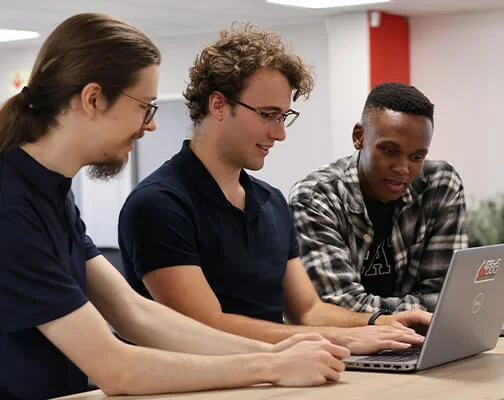 Three men collaborating at a table, engaged and smiling while looking at a laptop. The setting is a bright office with a red and white color scheme.