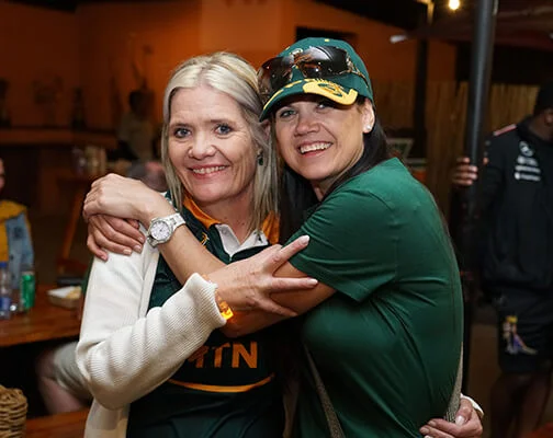 Two women in green sports shirts embrace and smile joyfully indoors. The atmosphere is casual, with warm lighting and people in the background.