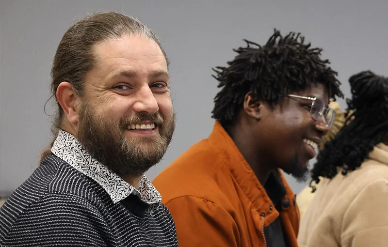 Two people sit and smile in a relaxed setting. The person on the left has a beard and wears a dark sweater; the person on the right wears glasses and an orange jacket.