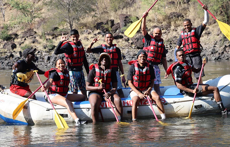 A group of eight smiling people in red life vests, holding yellow paddles, sit on a raft in a sunlit river, conveying adventure and joy.