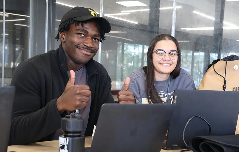 Two students sitting side by side at a table with laptops. Both are smiling and giving thumbs up gestures. The setting is a bright indoor space.