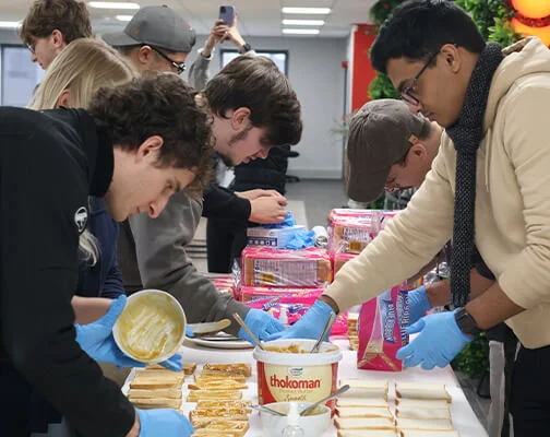 People preparing sandwiches at a table, spreading ingredients on bread. They wear gloves, focused on their task, creating a collaborative and caring atmosphere.