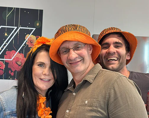 Three people smiling broadly; two wear orange hats, one with an orange flower headband. Bright, cheerful atmosphere with floral decor in the background.