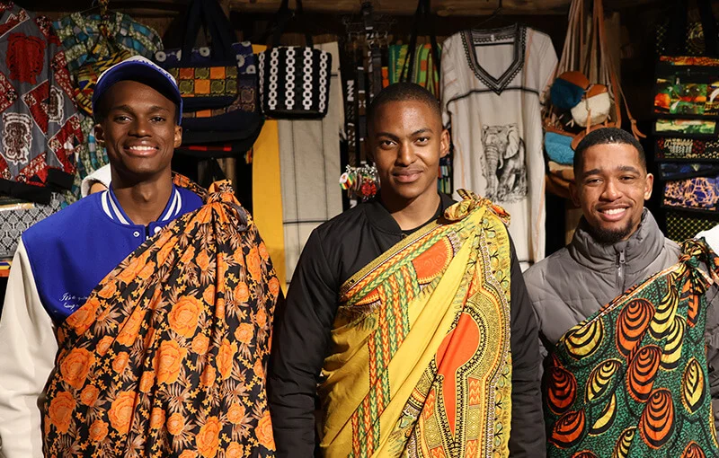 Three smiling men in brightly patterned shawls pose in a vibrant, colorful market stall. Background features an array of textiles and fabric.