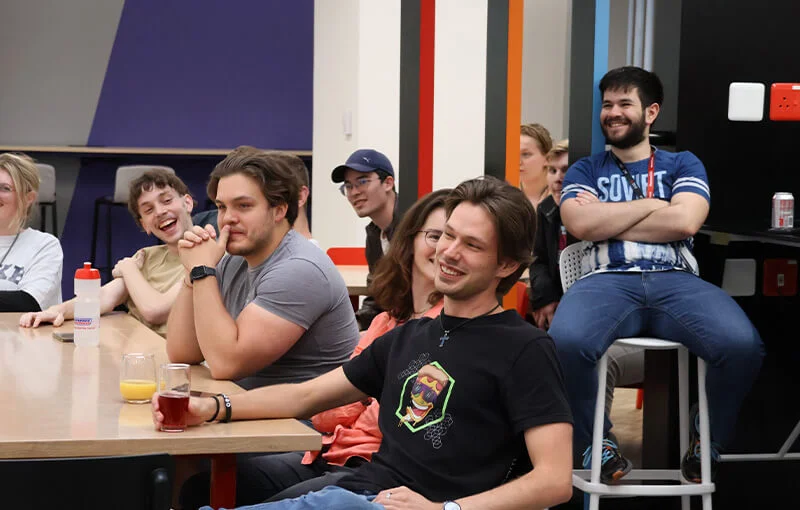 A group of people sit in a modern, colorful room, laughing and engaged. The mood is relaxed and cheerful, with several glasses on the table.