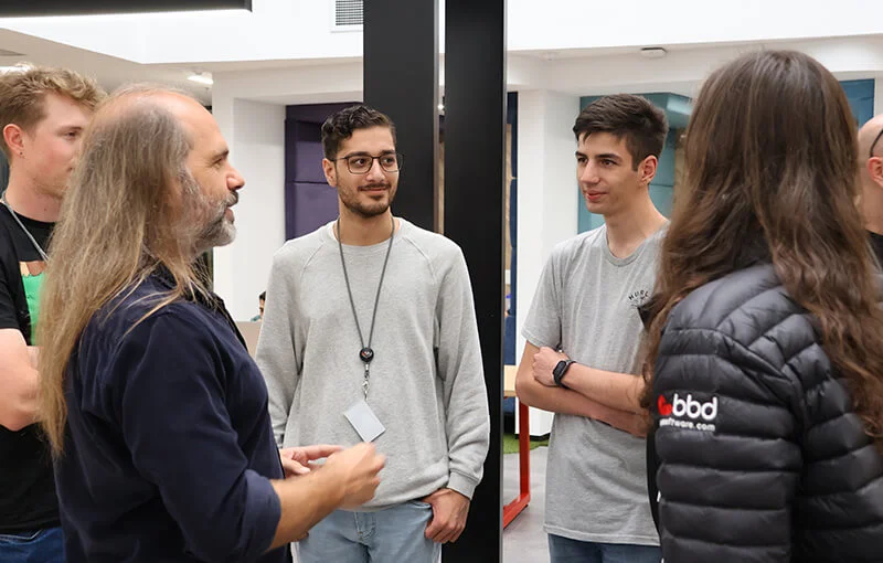 A group of five people, wearing casual clothes, are engaged in a lively conversation indoors. One person has long hair, creating a relaxed atmosphere.