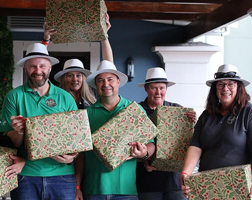 Five people in white hats hold Christmas-themed wrapped gifts, smiling cheerfully. They stand in a cozy, festive indoor setting.