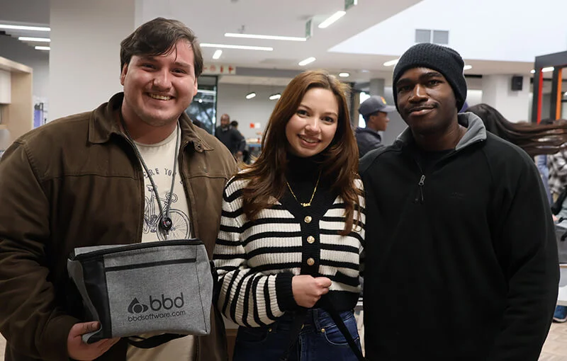 Three people are smiling indoors. The person on the left holds a gray bag. The atmosphere appears friendly and casual.