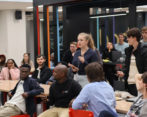 A woman speaks confidently in a modern office setting, surrounded by attentive colleagues, some seated and some standing, suggesting a collaborative discussion.