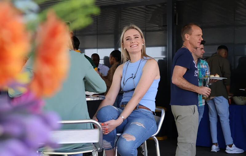 Woman in a blue sleeveless top and ripped jeans smiles, seated in a social gathering. Out-of-focus flowers in the foreground. Casual and cheerful tone.
