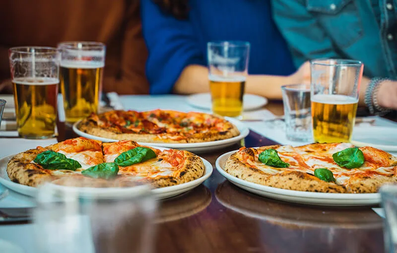 Three pizzas topped with basil leaves are on a table surrounded by three pints of beer and empty glasses. People are seated in the background.
