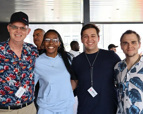 Four people stand closely smiling at an indoor event. They wear casual clothing with vibrant patterns. The mood is cheerful and welcoming.