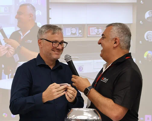 Two men are smiling as they engage in conversation. One holds a microphone while the other listens. A screen in the background displays the same scene.