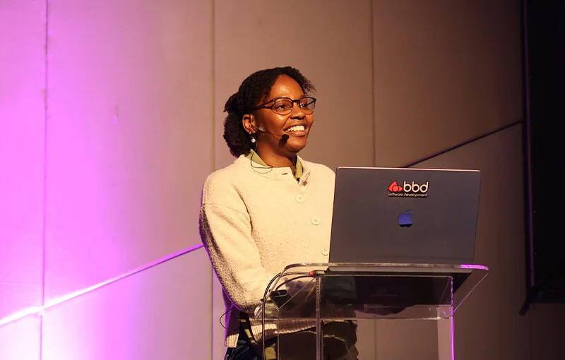A person is speaking confidently at a podium with a laptop. They are wearing glasses and beige attire. The background is lit with purple hues, creating a professional and engaging atmosphere.