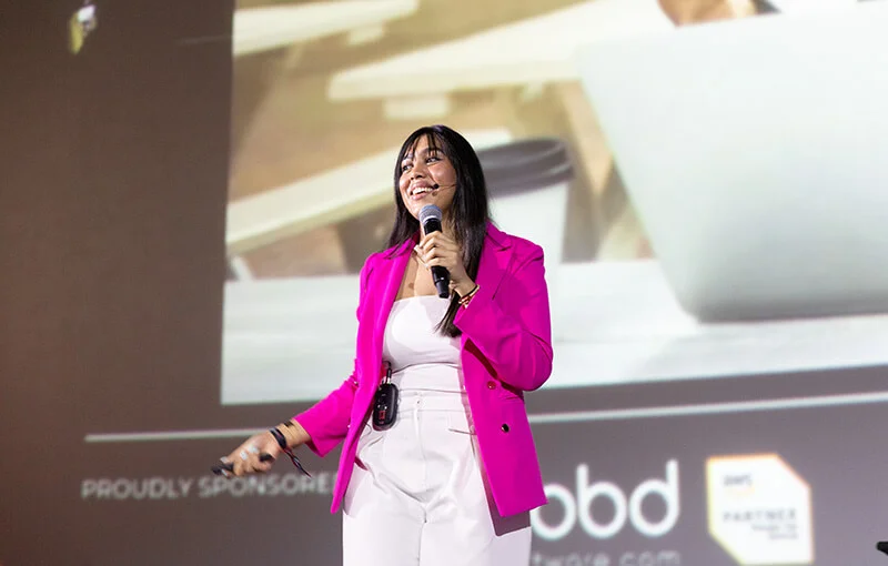 A woman in a bright pink blazer smiles and holds a microphone while speaking on stage. She stands in front of a large screen, exuding confidence and energy.
