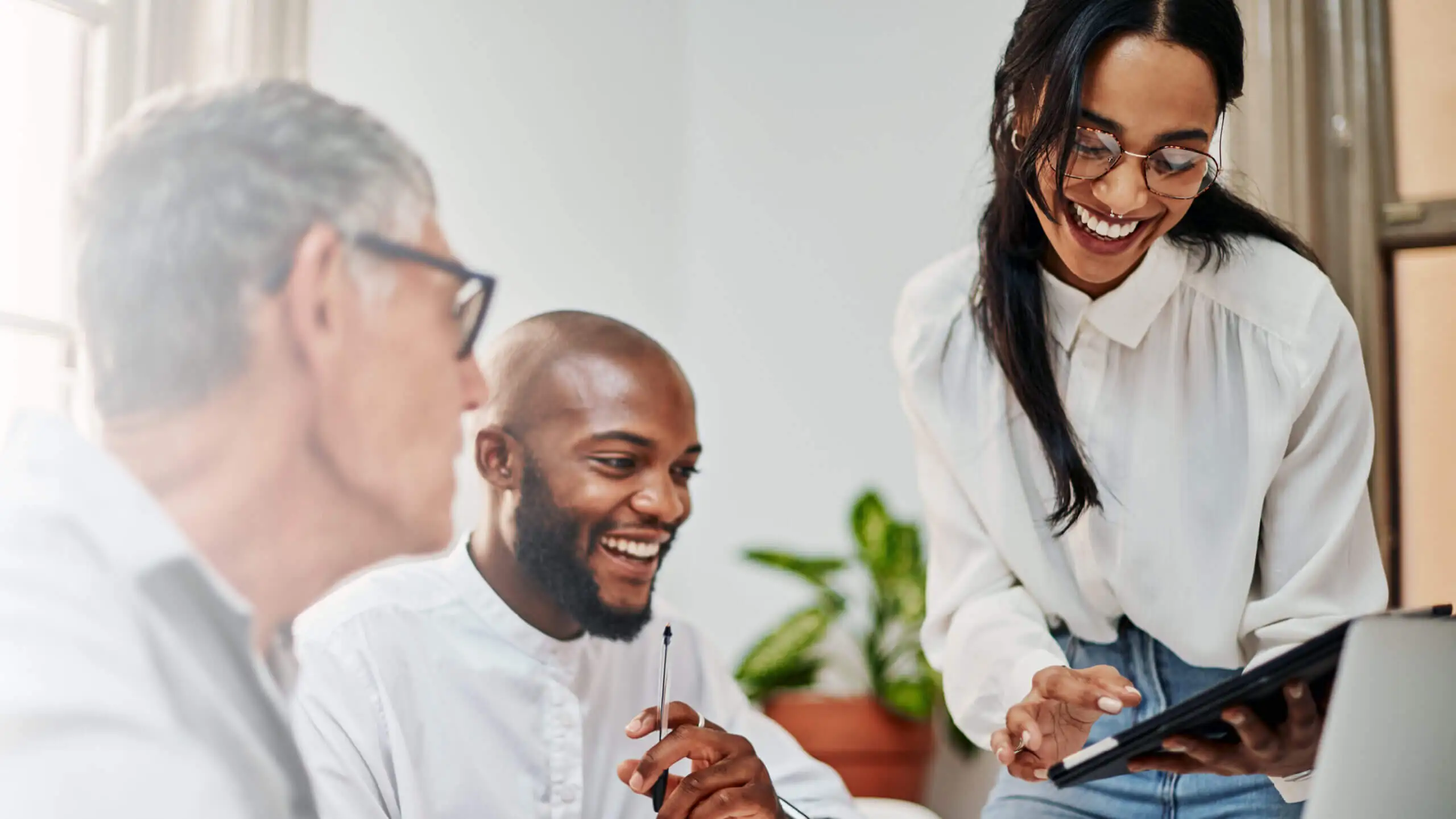 A diverse group of three colleagues engaged and smiling, gathered around a tablet. They're in a bright office space with natural light and a potted plant behind them.