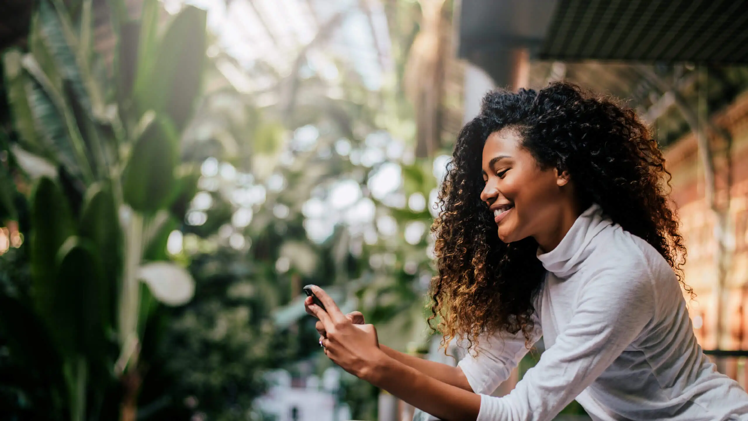 A woman with curly hair smiles while looking at her phone in a lush, green garden, sunlight filtering through