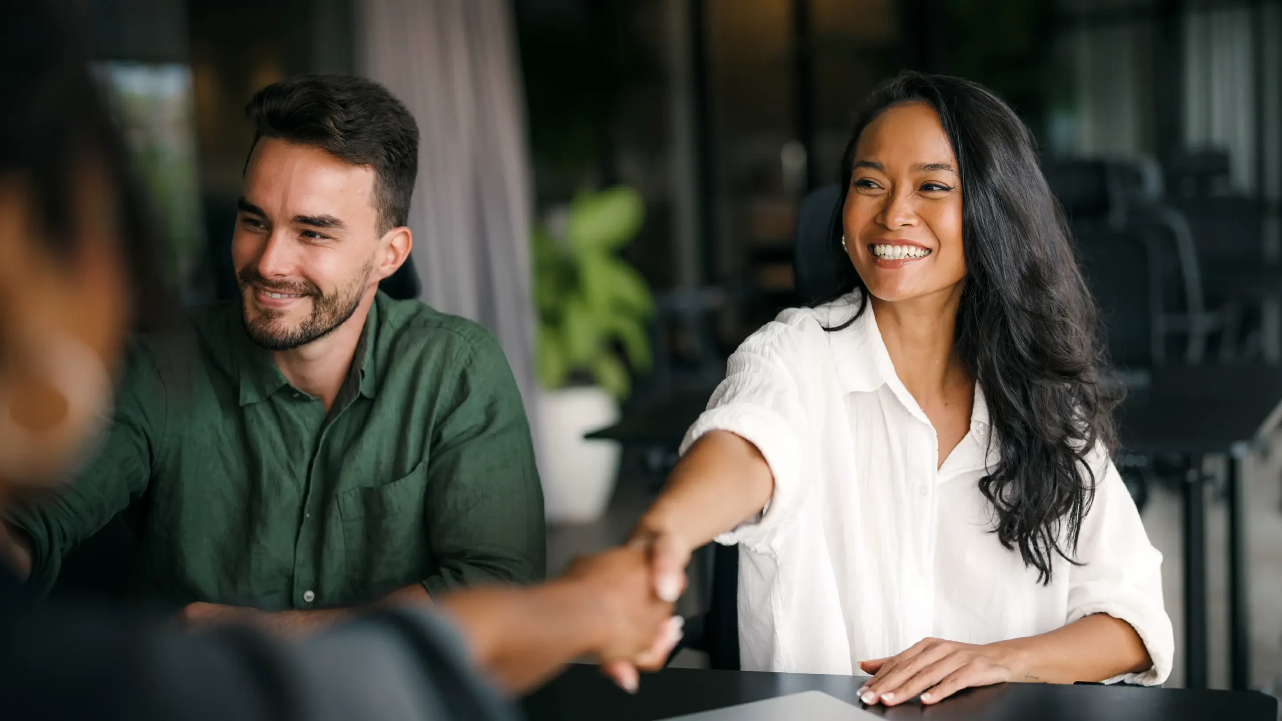 A woman in a white shirt smiles warmly while shaking hands across a table with another person. A man in a green shirt sits beside her, looking pleased.