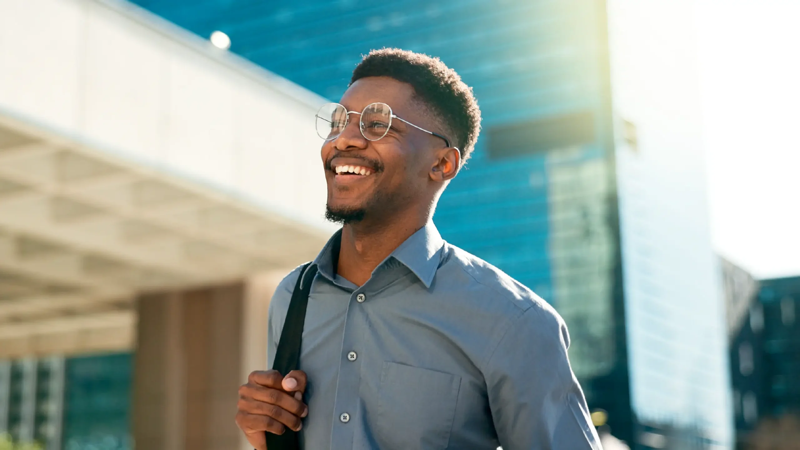 A smiling young professional with round glasses and a grey button-down shirt, walking outdoors in a bright city setting with modern glass buildings in the background.