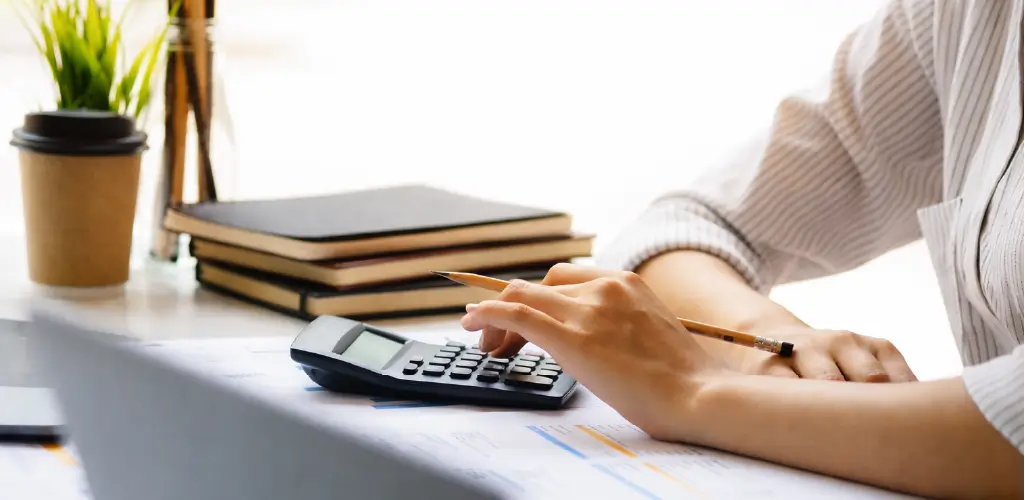 A person in a striped shirt uses a calculator on a desk with documents, a pencil, and books. A small plant and a coffee cup are nearby, conveying focus and productivity.