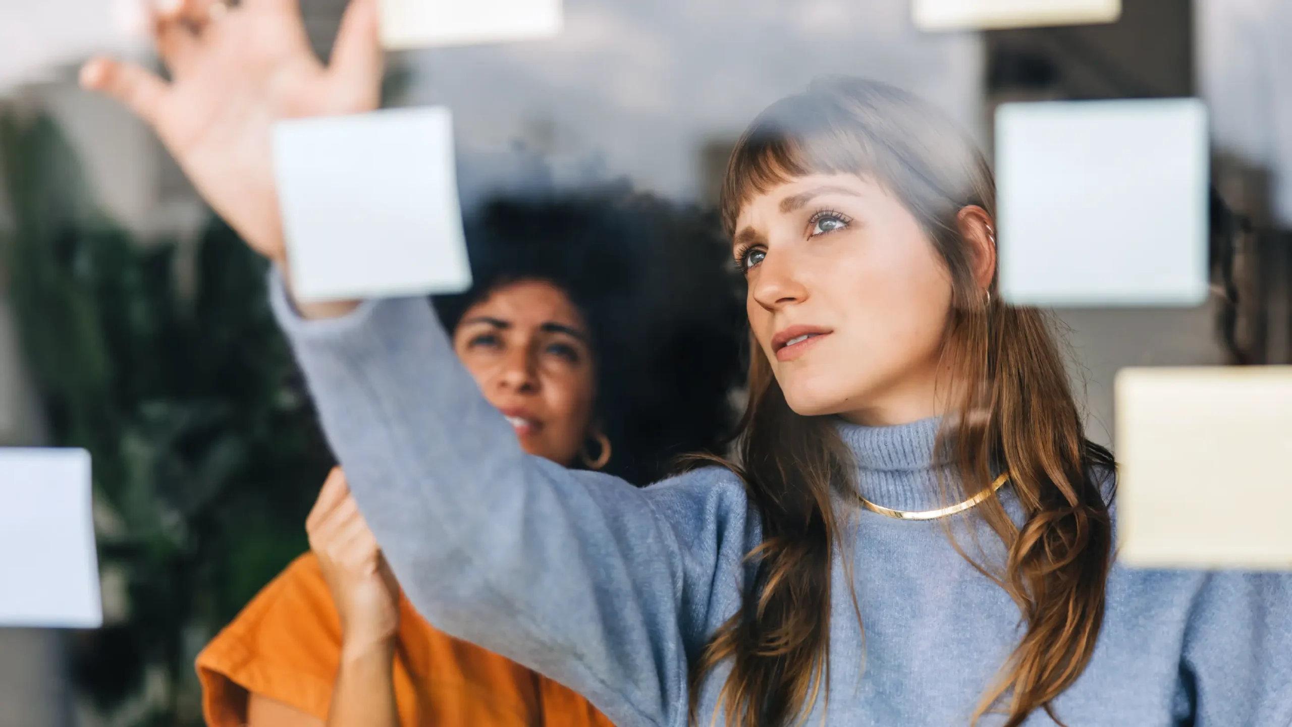 Two people seen through a glass wall with sticky notes attached, one in the foreground raising a hand toward the glass while the other stands behind.
