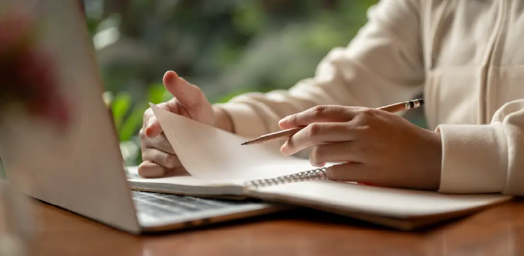 Close-up of hands writing in a notebook with a pencil, placed next to an open laptop on a wooden table.