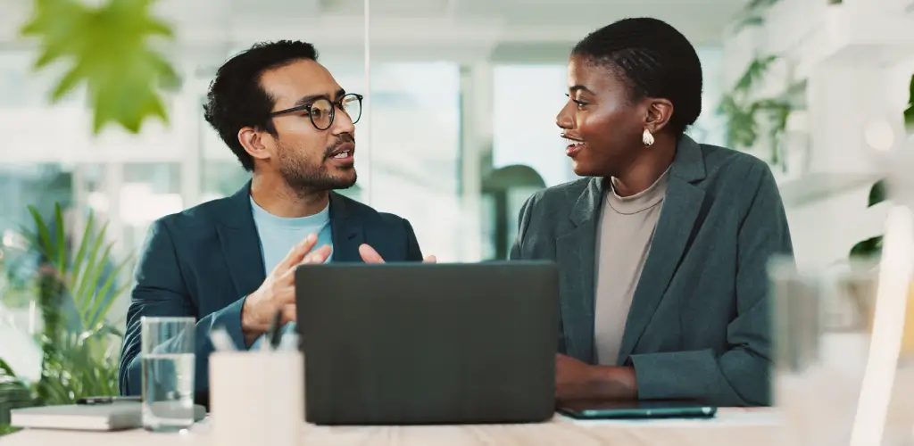 A man and woman in business attire converse animatedly at a desk, with a laptop open.