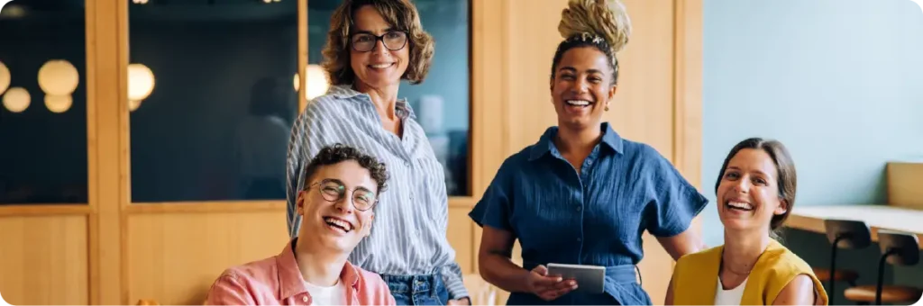 Four diverse colleagues smile and laugh together during a relaxed workplace gathering.