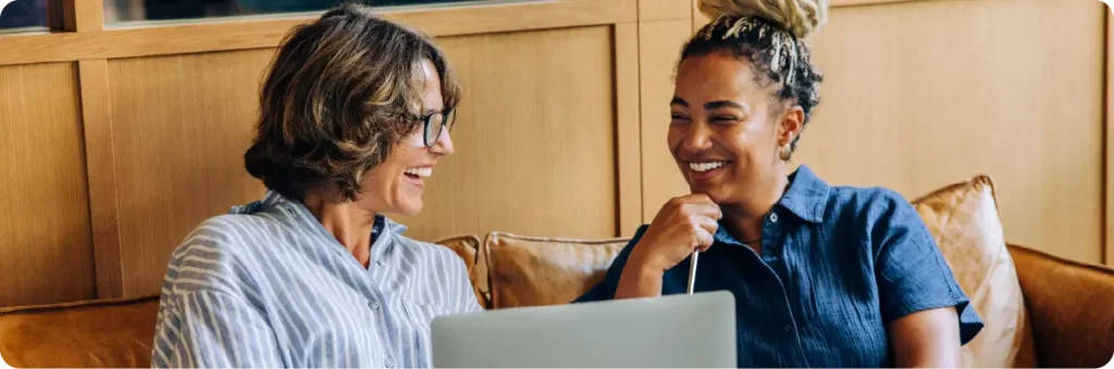 Two colleagues smile while collaborating on a laptop in a comfortable office seating area.