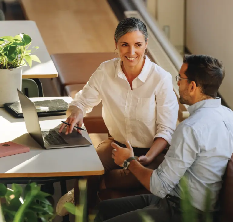 A smiling woman points to a laptop while discussing work with a seated male colleague in a bright office